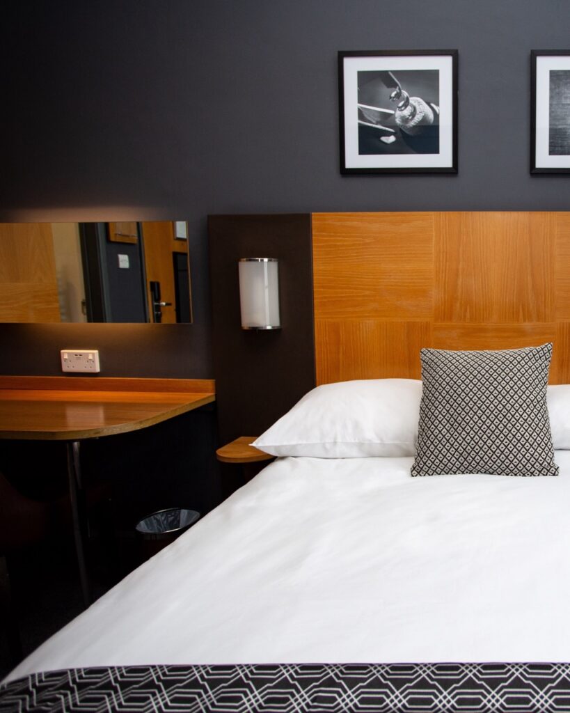 Hotel room scene: white bed with a patterned throw pillow, wooden headboard, dark wall, and a desk with a mirror on the left.